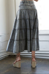 A model from behind wearing a high-waisted, tiered gray denim maxi skirt paired with brown espadrille wedge sandals in a bright room with white walls and a dark tiled floor.